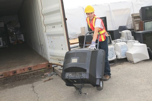 A worker wearing a yellow safety helmet, high-visibility vest with red and yellow reflective stripes, and work gloves is transporting an old, black computer monitor out of a white storage container or shipping unit. The monitor is a large, box-style CRT type with a curved screen and visible cables hanging from the back. The scene is set outdoors on a concrete surface, with the open container to the left revealing a dark interior with some shelving. Behind the worker, there are stacks of assorted electronic waste, including older desktop computers and other electronic devices, piled on the ground. The environment appears to be part of a waste management or rubbish removal operation, supporting services such as those offered by Commercial Waste Removal Norbiton, which specializes in disposing of electronic and general waste materials in the local area. The lighting suggests daytime conditions with natural light illuminating the scene clearly.