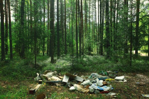 A scene of illegal rubbish dumping in a woodland area, featuring a mixture of discarded items such as cardboard boxes, plastic bags, and various waste materials scattered across the forest floor. The waste appears haphazardly piled on the ground amid tall, slender trees with green foliage, and the background shows a dense green forest with sunlight filtering through the canopy. The rubbish includes flattened cardboard cartons, soft plastic bags, and miscellaneous household waste, indicating an illegal waste disposal scene that commercial waste removal services in Norbiton, such as those offered by Commercial Waste Removal Norbiton, aim to address and remove from similar environments for proper clearance and cleanup.