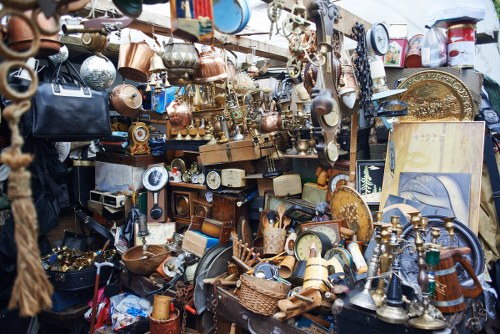 A densely packed collection of various discarded household items and decorative objects displayed in an outdoor setting, likely a market stall or area for rubbish collection in Norbiton. The assortment includes numerous brass and copper items such as teapots, trays, and candlestick holders with shiny, reflective surfaces, alongside wooden and ceramic objects like bowls, picture frames, and small boxes. Visible on the left are some fabric or rope materials, and in the background, other miscellaneous items are stacked or hung, creating a cluttered, chaotic appearance characteristic of a collection of waste or discarded goods. The lighting appears natural, highlighting the varied textures and finishes of the objects, from polished metals to matte ceramics. This scene relates to waste removal services offered by Commercial Waste Removal Norbiton, illustrating a typical example of bulky rubbish or clearance items that may require professional disposal, especially in urban or suburban environments. The setting is outdoor, on a street or pavement in a residential or commercial area, with a focus on the diverse materials and the disorganized nature of the unwanted items ready for collection or disposal in the local borough.