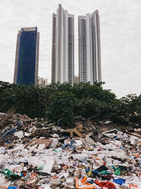 A large pile of mixed household and building waste, including plastic bags, cardboard boxes, foam pieces, and discarded debris, is spread across the ground in the foreground of the image. Behind the w