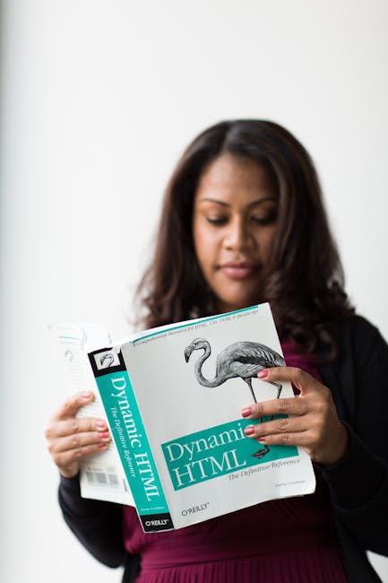 A woman with dark, shoulder-length hair is standing indoors against a plain light-colored wall. She is holding an open book titled 'Dynamic HTML' with a large illustration of a flamingo on the cover, 
